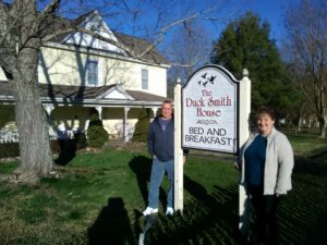 Marcia & Mark stand in front of the bed & breakfast sign.